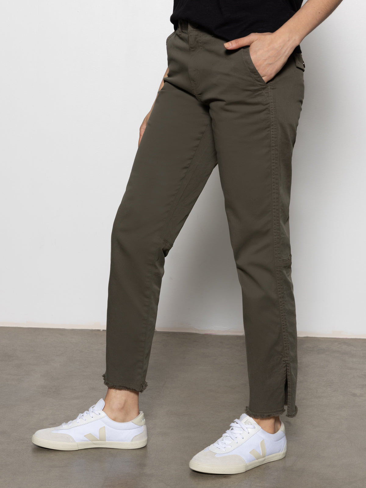 A person wearing Sanctuary Clothing's "Trade Maker Pant" in kalamata and white sneakers stands casually on a gray floor, with one hand in their pocket. They're also wearing a black top, and the photo is cropped just above the waist.