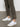 A person in Sanctuary Clothing's trade maker pant kalamata, featuring frayed hems and side slits, stands on a concrete floor. They pair it with white sneakers that have beige soles and a white "V" logo.