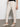 A person in Sanctuary Clothing's trade maker pant light oat and a black top stands against a white wall, one hand in their pocket. The pants feature small side slits, complementing their woven cream-colored shoes.