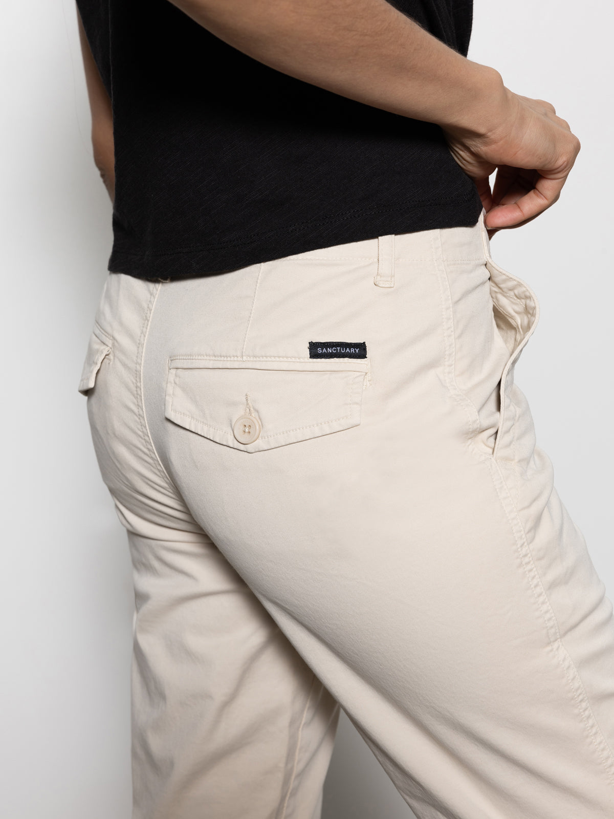 A person wearing Sanctuary Clothing's trade maker pant in light oat, featuring a visible brand label on the back pocket, paired with a black shirt, with their hand casually placed in the front pocket against a plain white background.