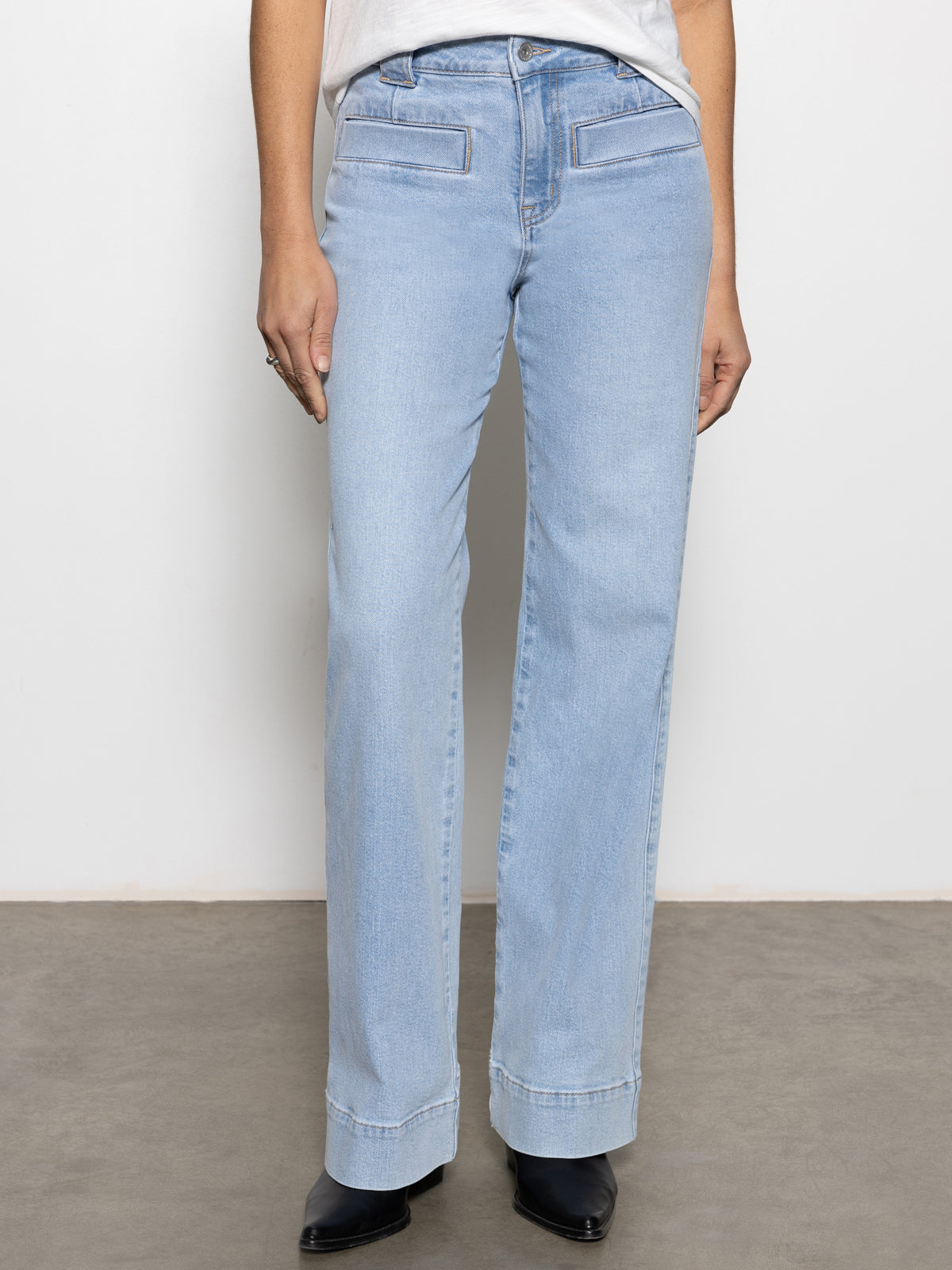 A person in Sanctuary Clothing's "voyage full length pant wishful thinking" light blue jeans with front flap pockets and a white shirt stands against a plain white wall. They are wearing black shoes, with the focus on the jeans from the waist down.