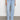 A person in Sanctuary Clothing's "voyage full length pant wishful thinking" light blue jeans with front flap pockets and a white shirt stands against a plain white wall. They are wearing black shoes, with the focus on the jeans from the waist down.