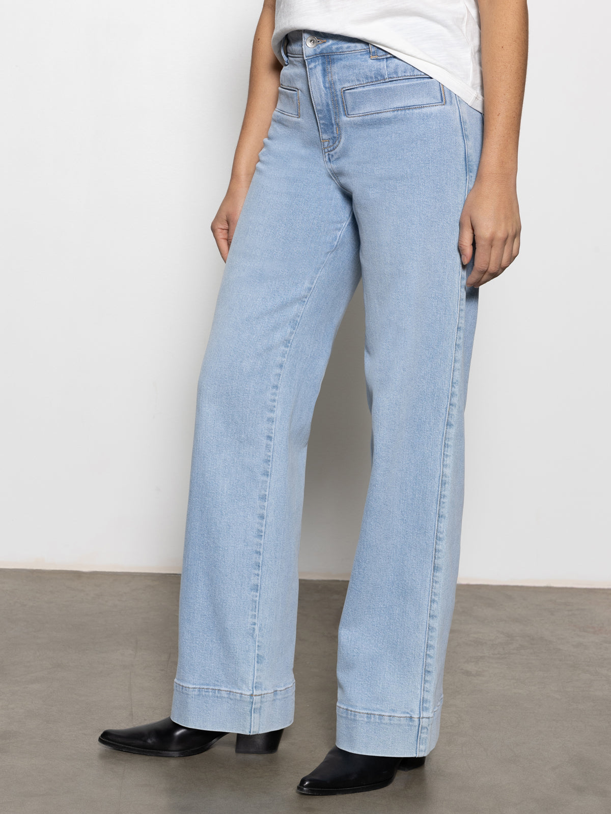 A person is wearing Sanctuary Clothing's Voyage Full-Length Pant in Wishful Thinking—a pair of light blue high-waisted wide-leg jeans—and a white top, standing against a plain white background. They complete the look with black ankle boots.