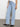 A person is wearing Sanctuary Clothing's Voyage Full-Length Pant in Wishful Thinking—a pair of light blue high-waisted wide-leg jeans—and a white top, standing against a plain white background. They complete the look with black ankle boots.