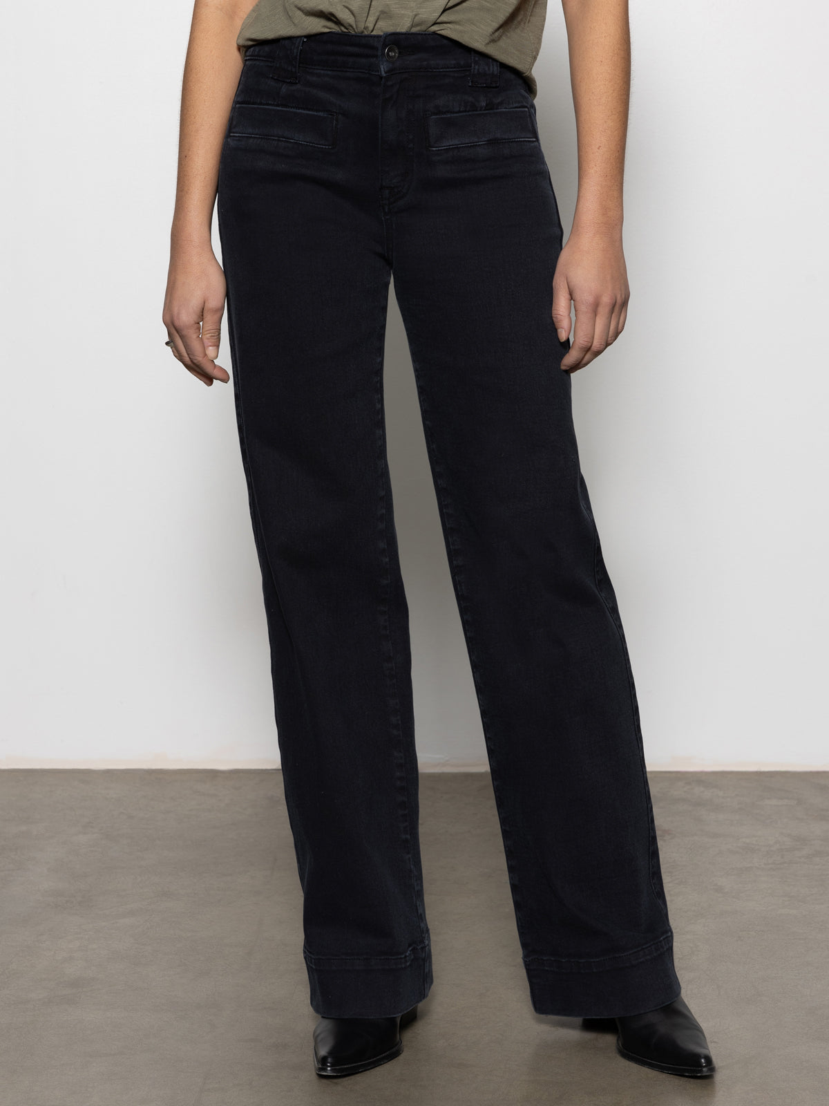 A person stands against a plain white wall on a concrete floor, wearing Sanctuary Clothing's Voyage full-length wide-leg black jeans paired with a dark green top and black pointed-toe shoes. The jeans feature a high waist design.