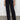 A person stands against a plain white wall on a concrete floor, wearing Sanctuary Clothing's Voyage full-length wide-leg black jeans paired with a dark green top and black pointed-toe shoes. The jeans feature a high waist design.