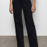 A person stands against a plain white wall on a concrete floor, wearing Sanctuary Clothing's Voyage full-length wide-leg black jeans paired with a dark green top and black pointed-toe shoes. The jeans feature a high waist design.