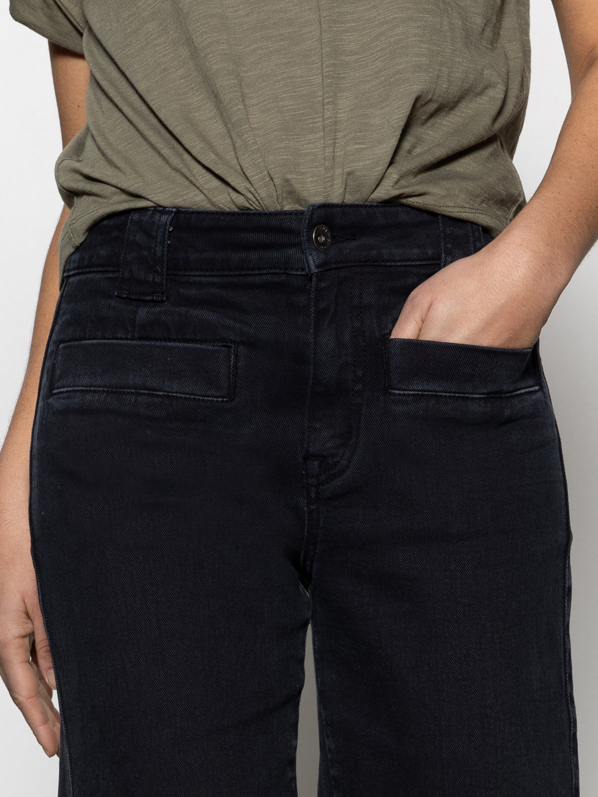 A person wearing Sanctuary Clothing's voyage full length black jeans and a green t-shirt has their right thumb tucked into the pocket, with the t-shirt fabric slightly bunched at the waist.