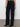 A person is standing against a light wall and gray floor, wearing Sanctuary Clothing's voyage full-length black jeans, featuring front pockets and high-waisted, straight legs. They're paired with a tucked-in grayish shirt and black heeled ankle boots.