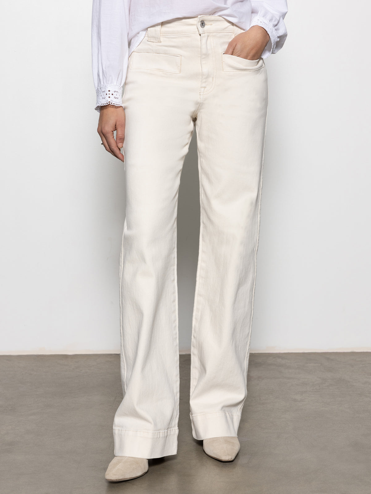 Someone wearing Sanctuary Clothing's "Voyage Full Length Pant" in light oat, with one hand in the pocket, paired with a white blouse featuring lace-detailed cuffs and beige pointed shoes, is standing against a plain background.