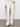 Someone wearing Sanctuary Clothing's "Voyage Full Length Pant" in light oat, with one hand in the pocket, paired with a white blouse featuring lace-detailed cuffs and beige pointed shoes, is standing against a plain background.