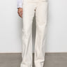 Someone wearing Sanctuary Clothing's "Voyage Full Length Pant" in light oat, with one hand in the pocket, paired with a white blouse featuring lace-detailed cuffs and beige pointed shoes, is standing against a plain background.