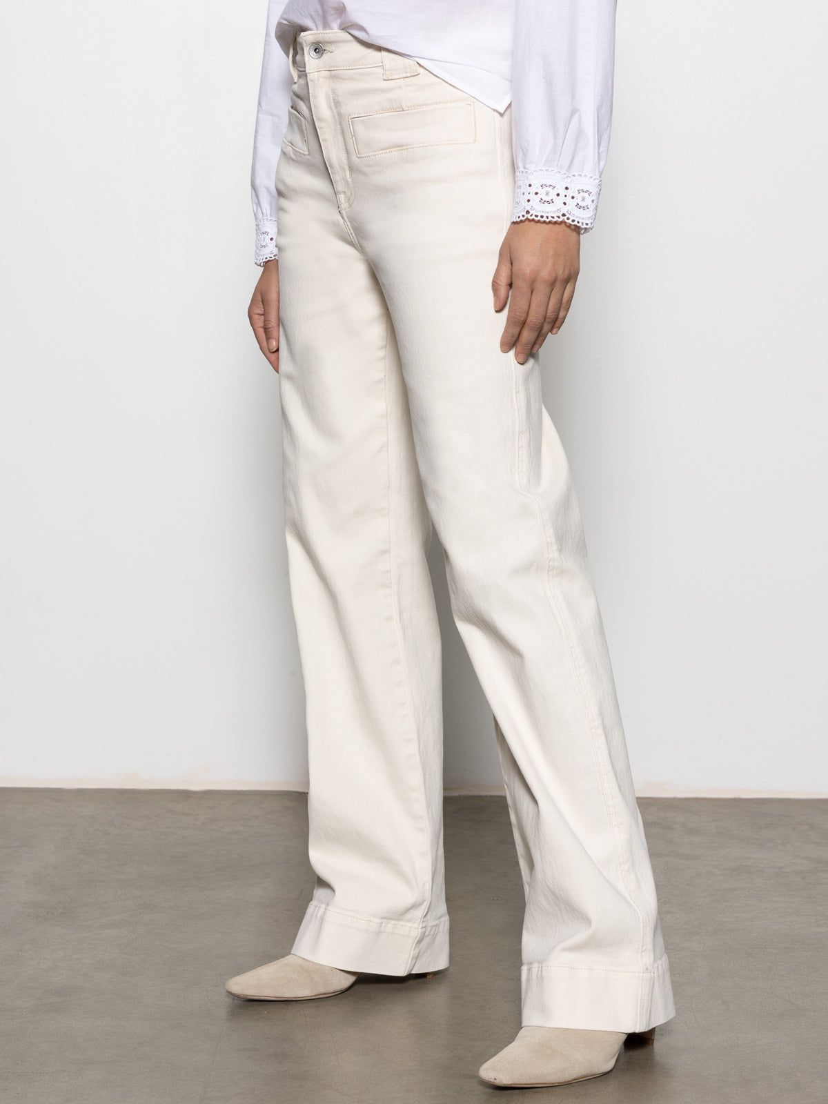 A person stands on a concrete floor wearing a white long-sleeve shirt with lace cuffs and Sanctuary Clothing's "voyage full length pant" in light oat, complemented by beige shoes, against a plain white wall.