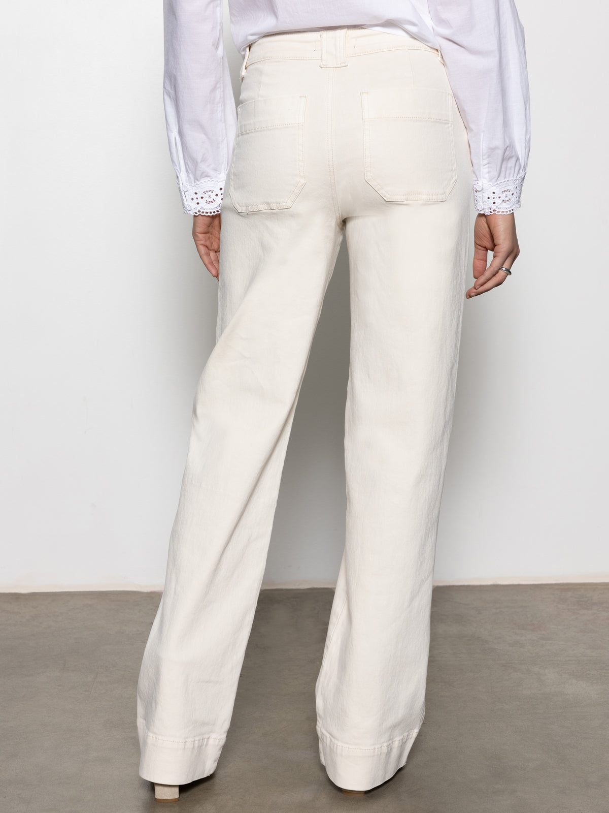 Rear view of a person wearing Sanctuary Clothing's Voyage Full Length Pant in light oat, paired with a white blouse featuring eyelet sleeve details. They stand on a gray floor against a neutral background.