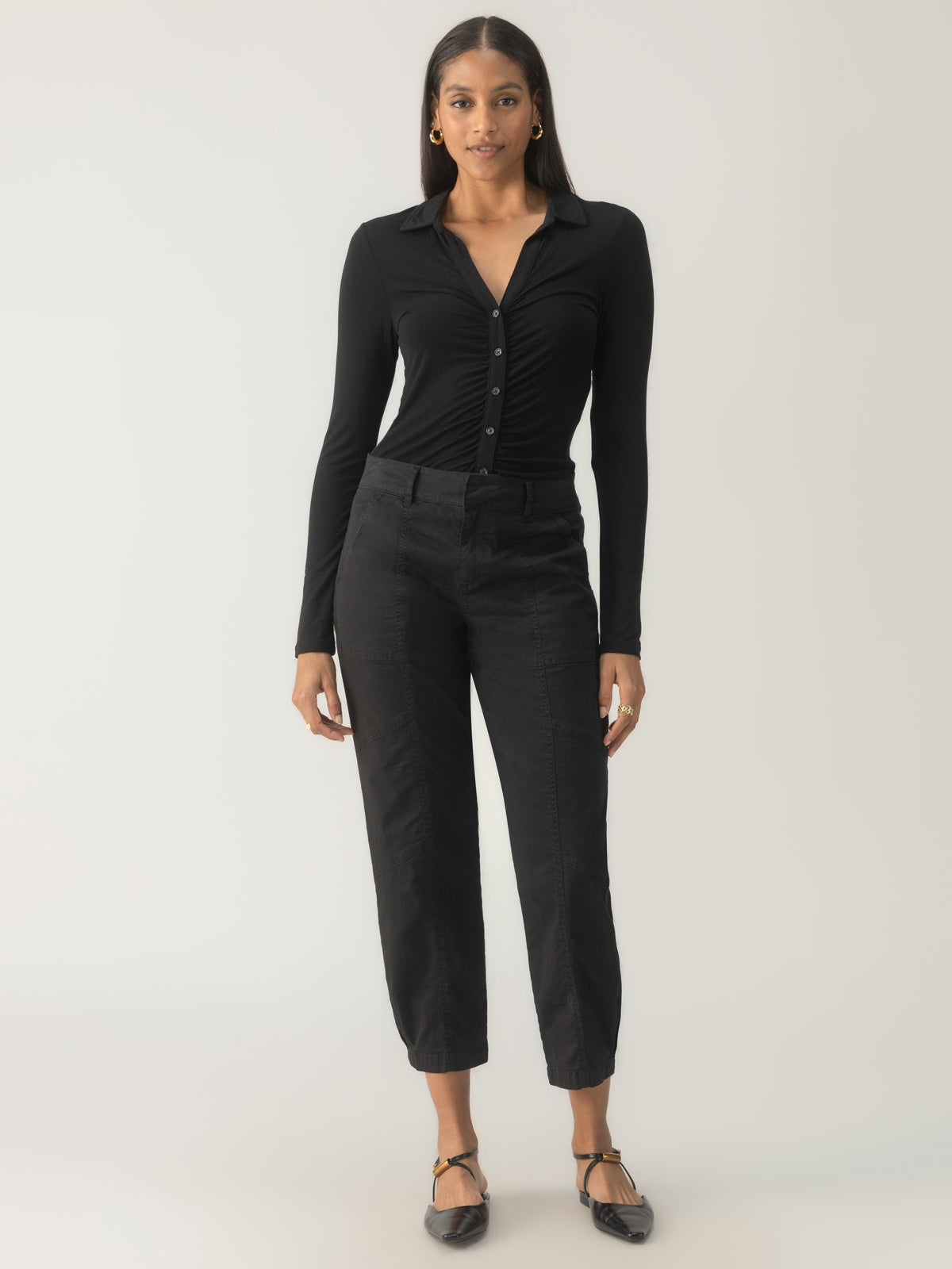 A person stands against a neutral background wearing the Sahara Tapered Pant in black from Sanctuary Clothing. They are also dressed in a black long-sleeve buttoned shirt, complemented by black flats and gold hoop earrings, creating a sleek and monochromatic outfit.