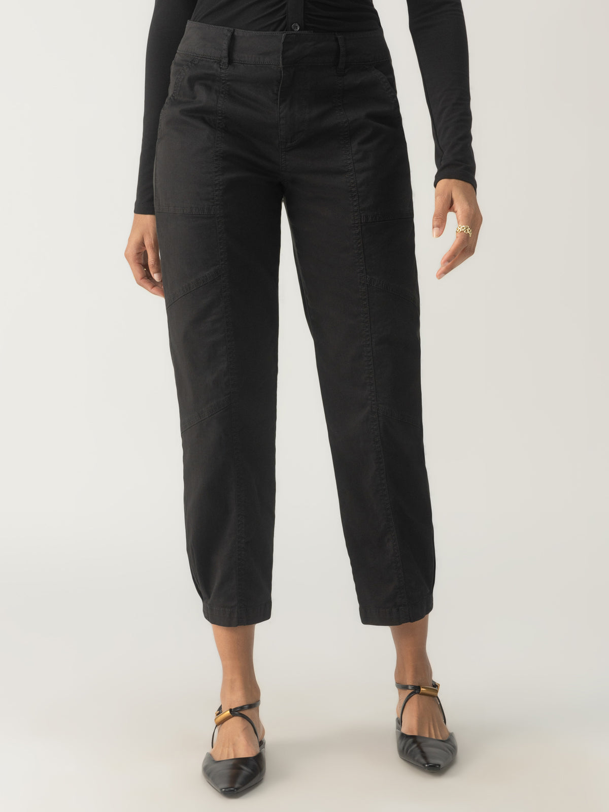 A person is wearing the "Sahara Tapered Pant Black" by Sanctuary Clothing, which features a relaxed fit and cropped length, complete with convenient pockets. They have paired it with a black long-sleeve top and stylish black pointed-toe shoes with ankle straps. The backdrop is a plain white setting.