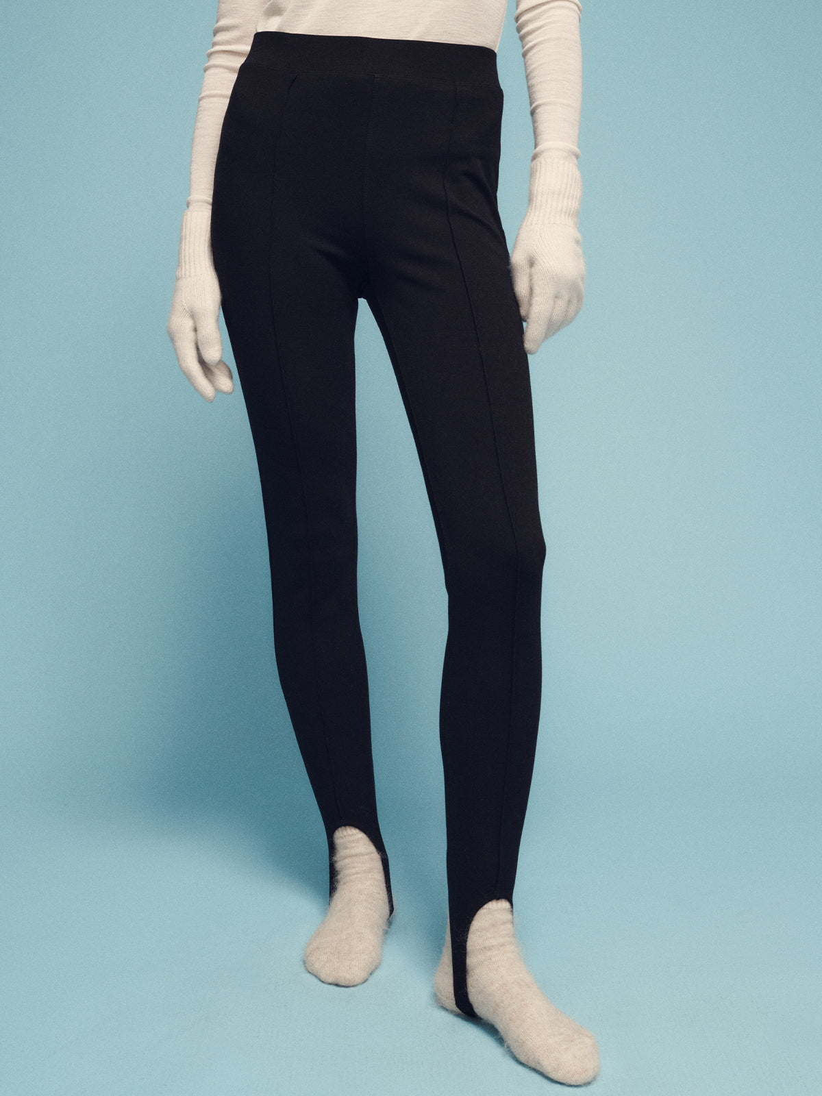 A person wearing a long-sleeved white top, stirrup legging black by Sanctuary Clothing, white gloves, and white socks stands against a light blue background. The image focuses on their legs and waist.