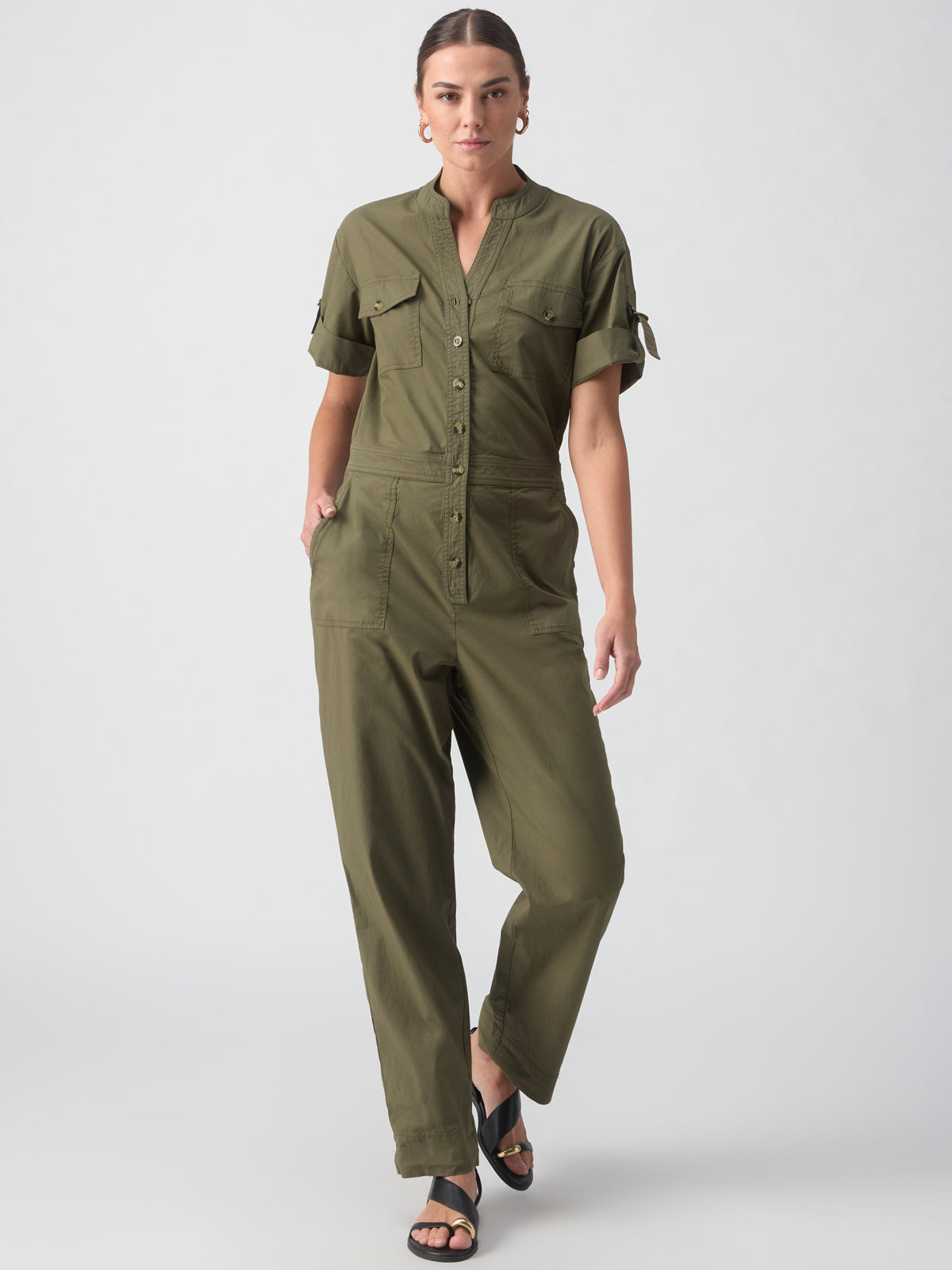 A woman stands against a plain gray background, wearing the Reserve Jumpsuit in Burnt Olive by Sanctuary Clothing. The jumpsuit features a button-up front, short sleeves, and pockets. She has minimal jewelry, including small hoop earrings, and wears black open-toe sandals. Her hair is pulled back in a low bun.