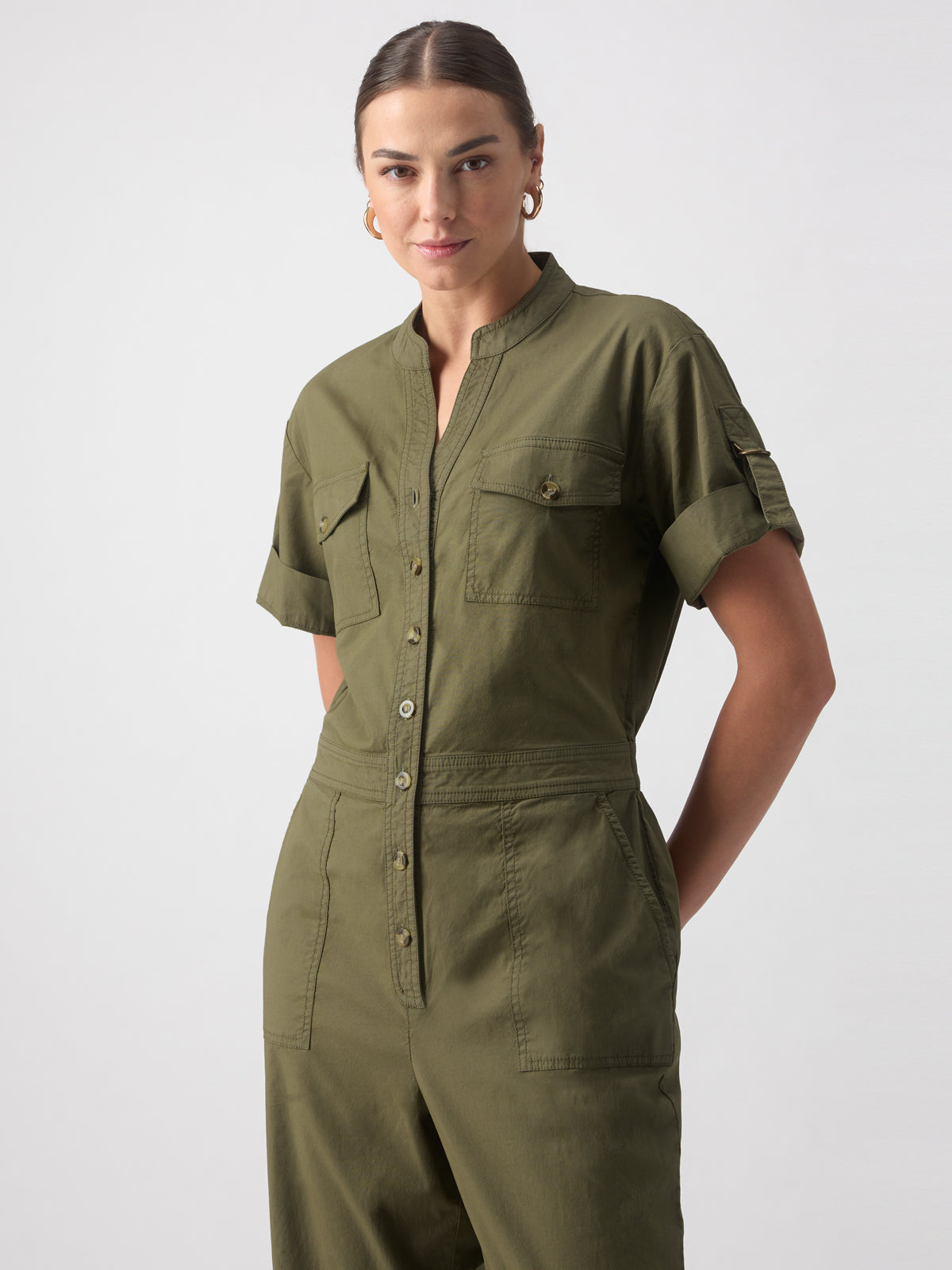 A person with their hair tied back is wearing the Reserve Jumpsuit Burnt Olive by Sanctuary Clothing, featuring short sleeves, a button-down front, and two chest pockets. The individual is standing against a plain light gray background and looking directly at the camera with a slight smile.