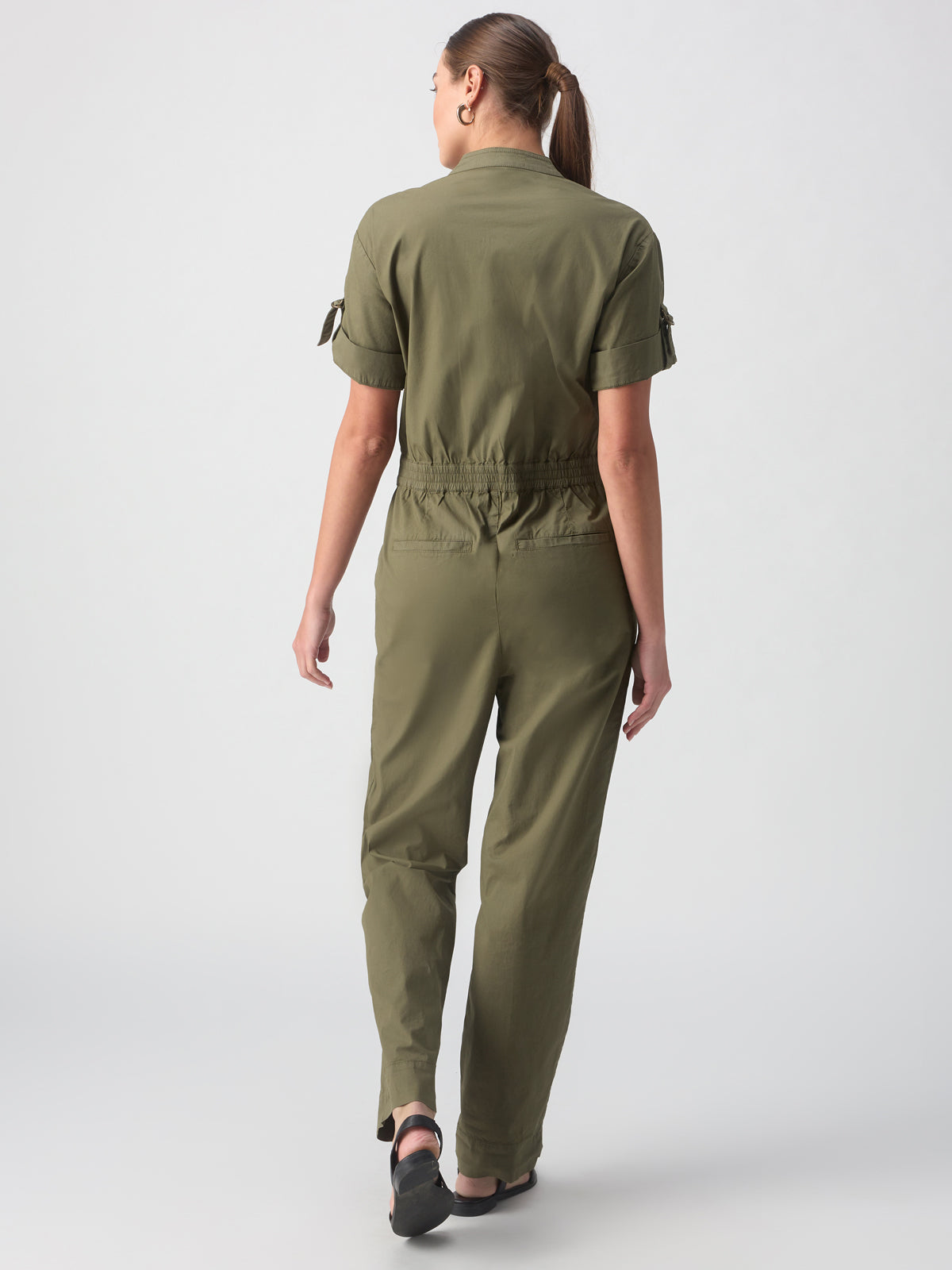 A person with a ponytail is standing facing away from the camera. They are wearing the Reserve Jumpsuit in Burnt Olive by Sanctuary Clothing, paired with black shoes. The background is plain and light-colored.