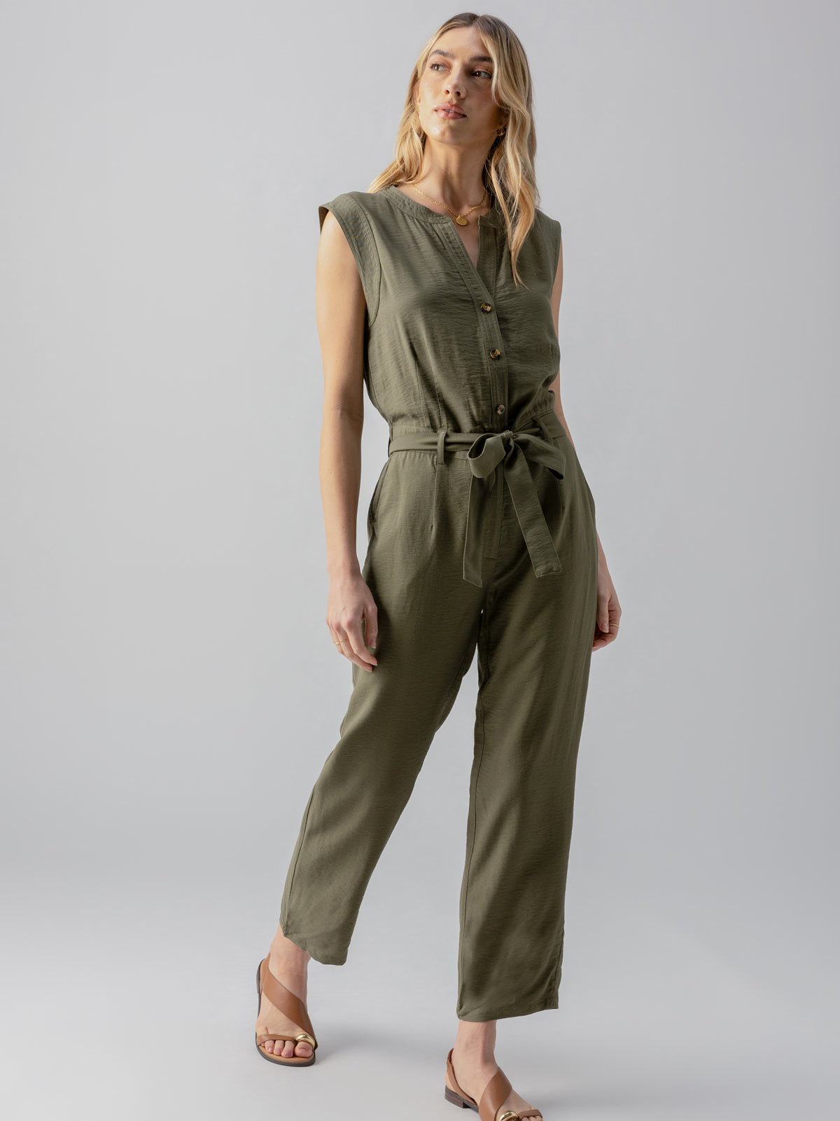 A person with long hair is wearing the Prima Jumpsuit in Burnt Olive by Sanctuary Clothing, which is sleeveless and tied at the waist. They are standing against a plain, light gray background with their head slightly tilted. They are also wearing tan sandals.