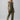 A person with long hair is wearing the Prima Jumpsuit in Burnt Olive by Sanctuary Clothing, which is sleeveless and tied at the waist. They are standing against a plain, light gray background with their head slightly tilted. They are also wearing tan sandals.