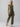 A person with long hair is wearing the Prima Jumpsuit in Burnt Olive by Sanctuary Clothing, which is sleeveless and tied at the waist. They are standing against a plain, light gray background with their head slightly tilted. They are also wearing tan sandals.