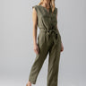 A person with long hair is wearing the Prima Jumpsuit in Burnt Olive by Sanctuary Clothing, which is sleeveless and tied at the waist. They are standing against a plain, light gray background with their head slightly tilted. They are also wearing tan sandals.