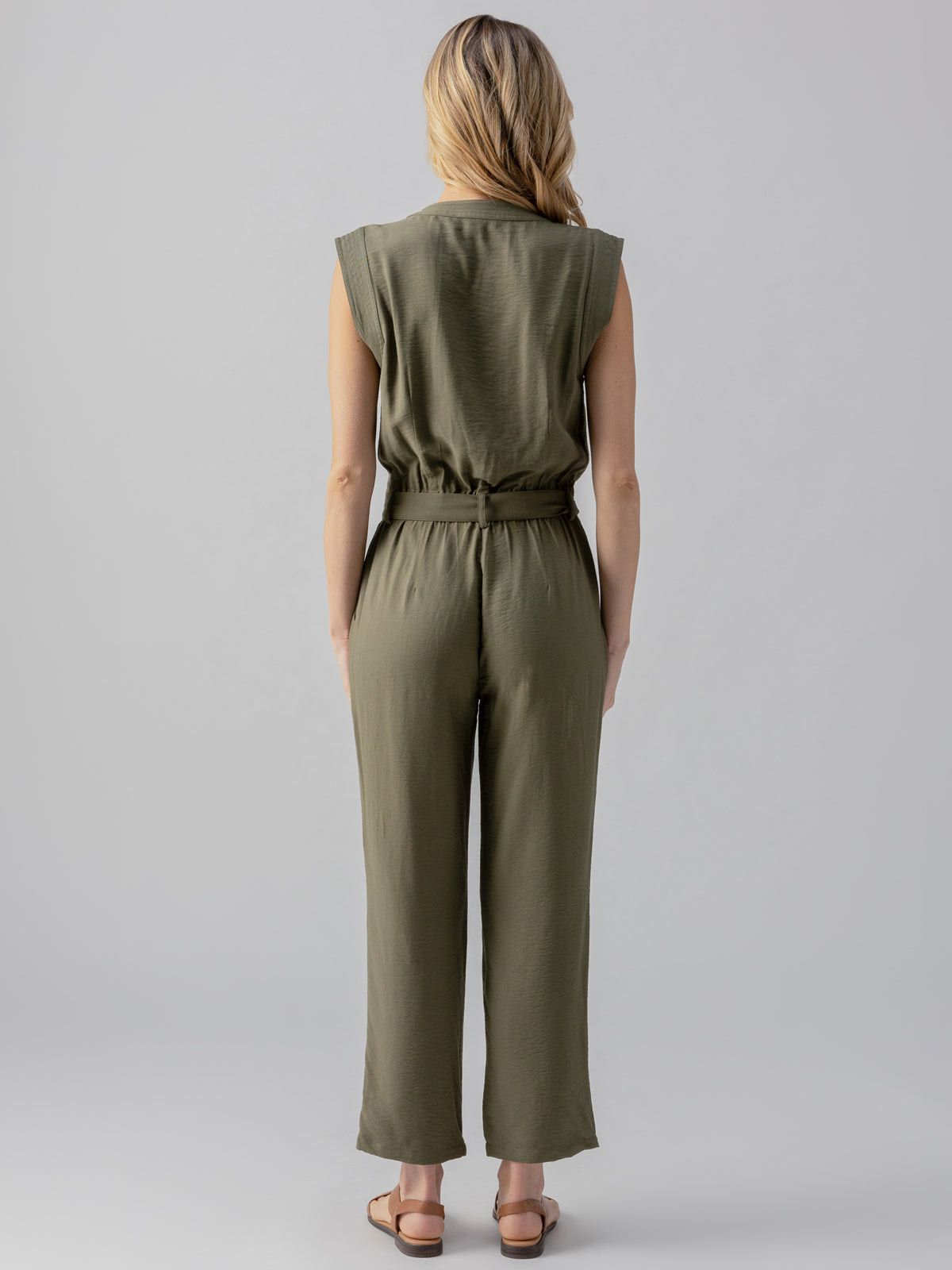 A person with long, wavy hair is facing away from the camera. They are wearing the Prima Jumpsuit in Burnt Olive by Sanctuary Clothing, which features a sleeveless design and a belt at the waist. The person is standing against a plain, light grey background and is wearing flat, brown sandals.