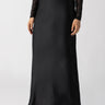 A person stands against a plain background, wearing the Sanctuary Clothing Everyday Maxi Skirt in black and a black lace long-sleeved top. They are also wearing pointed black flats and have one hand with a ring, resting by their side. The head is not visible in the image.