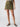 A person is wearing the Surplus Essential Skirt Burnt Olive from Sanctuary Clothing, a knee-length cargo skirt featuring two large front pockets. They are also wearing a striped top and black sandals. The photo is taken in a well-lit studio with a plain background.