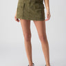 A person is wearing the Surplus Essential Skirt Burnt Olive from Sanctuary Clothing, a knee-length cargo skirt featuring two large front pockets. They are also wearing a striped top and black sandals. The photo is taken in a well-lit studio with a plain background.