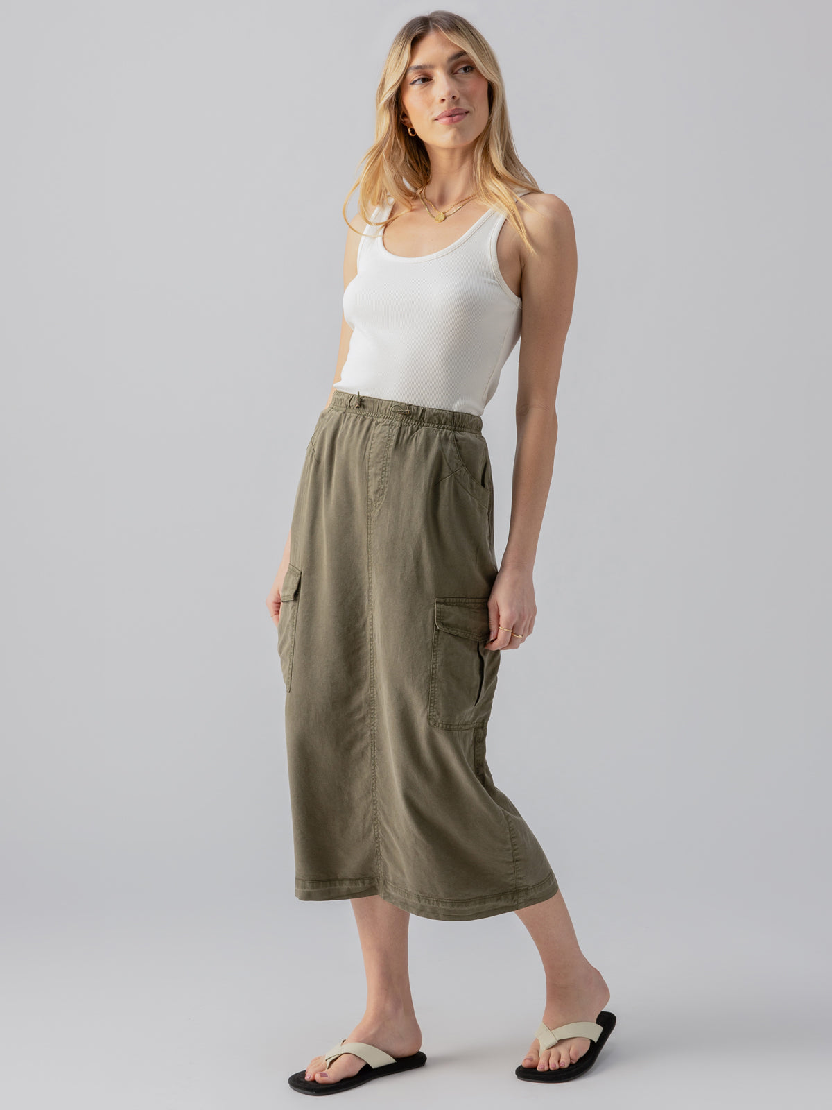 A woman with long, blonde hair stands against a plain background. She is wearing a white tank top, the Parachute Skirt Burnt Olive by Sanctuary Clothing, complete with pockets, and black sandals. She is looking slightly off to the side and posing with one hand on her hip.