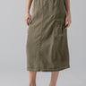 A person is wearing the Parachute Skirt Burnt Olive by Sanctuary Clothing, a knee-length cargo skirt with an elastic waistband and large side pockets. They are also dressed in a white tank top and black sandals with light-colored straps. The background is plain light gray.