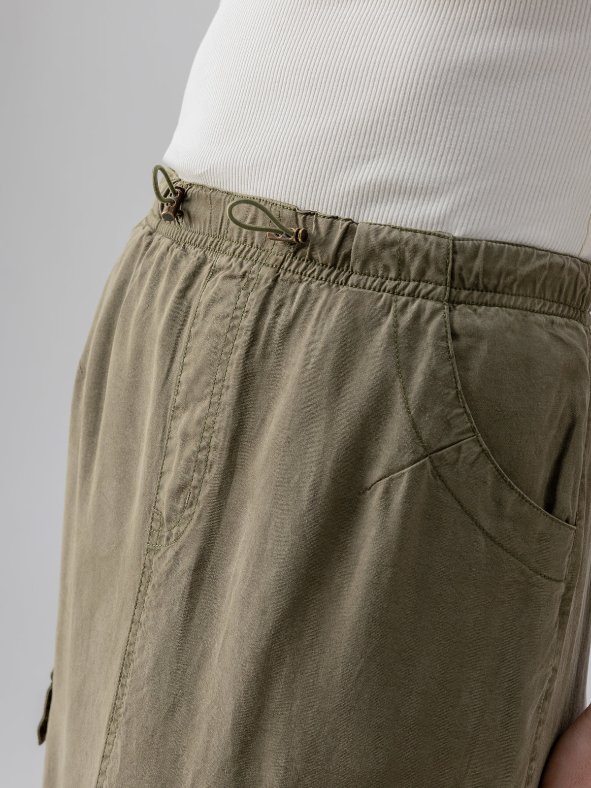 Side view of a person wearing the Sanctuary Clothing Parachute Skirt Burnt Olive with an elastic drawstring waist and a white ribbed top. The skirt features visible pockets, seams, and stitching details. The background is plain and neutral in color.