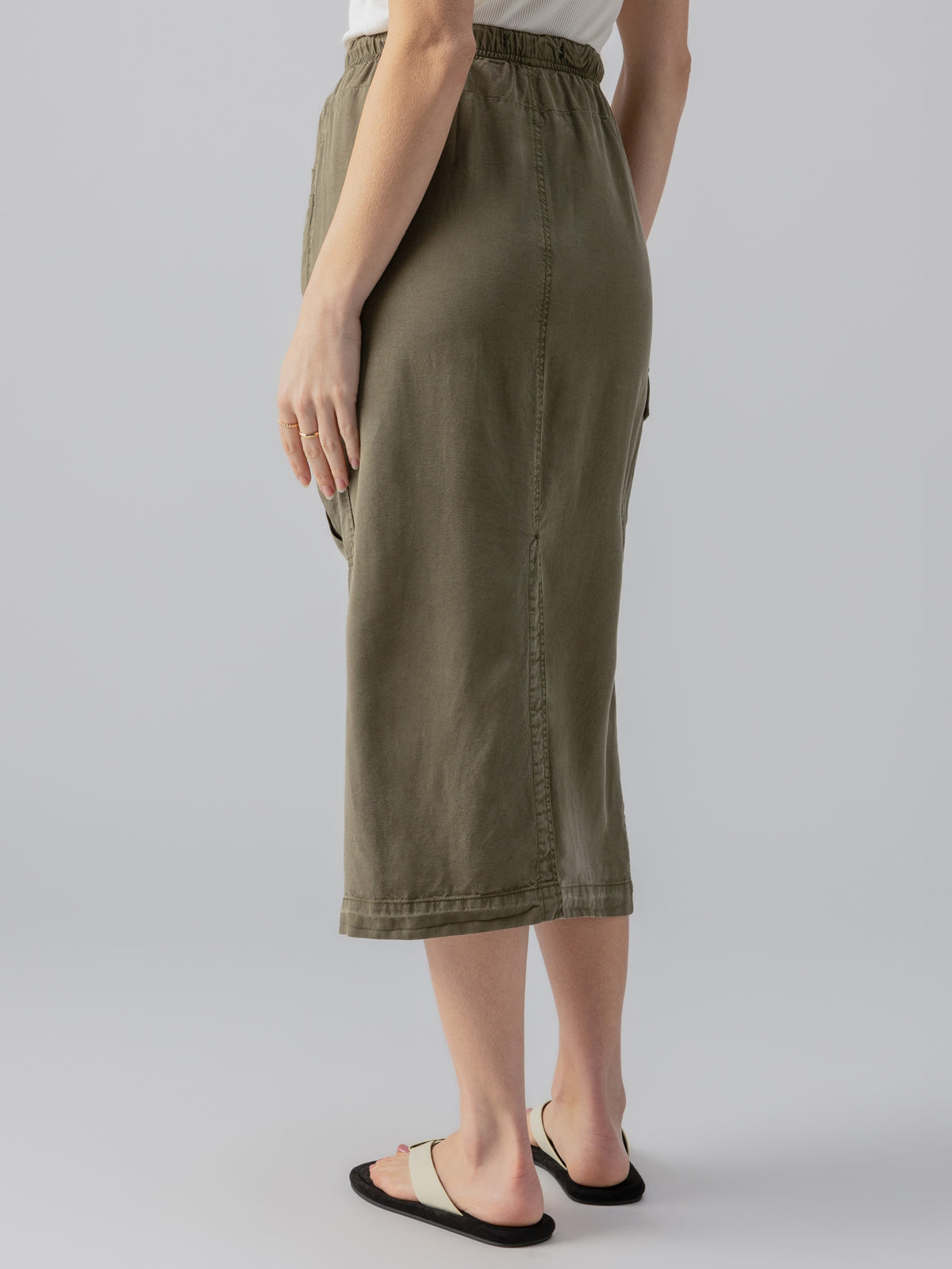 A person is standing with their back to the camera, wearing the Parachute Skirt Burnt Olive from Sanctuary Clothing. The skirt is midi-length with an elastic waistband. They are also wearing a white top and beige slide sandals with black soles. The background is plain and light gray.