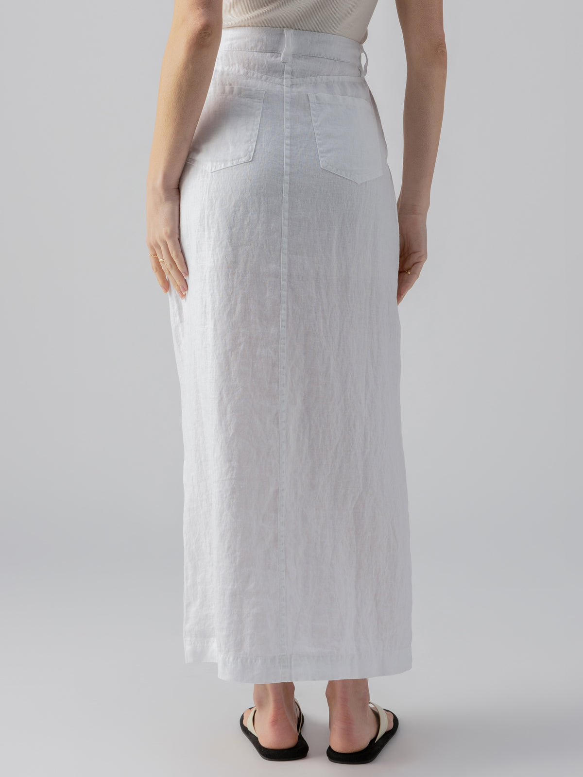 A person is shown from the back wearing the Boardwalk Slip Skirt White by Sanctuary Clothing, a high-waisted, ankle-length white linen skirt with back patch pockets. They are also sporting a white top and black sandals against a plain, light gray background.