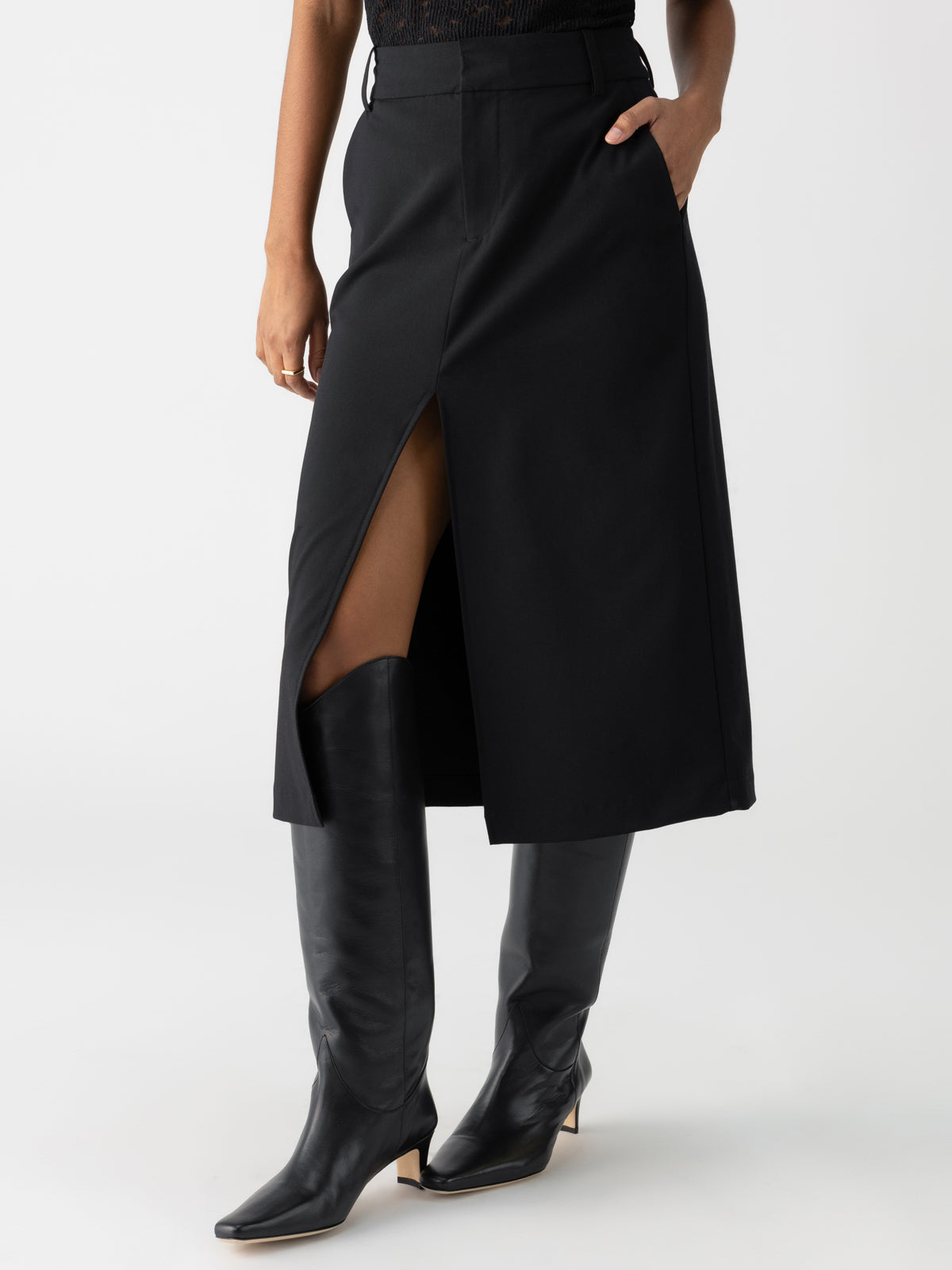 A person wearing the Classic Slit Skirt Black by Sanctuary Clothing, featuring a front slit and convenient pockets, paired with matching black knee-high boots. The individual is striking a pose with one hand tucked into a pocket, set against a plain white background.