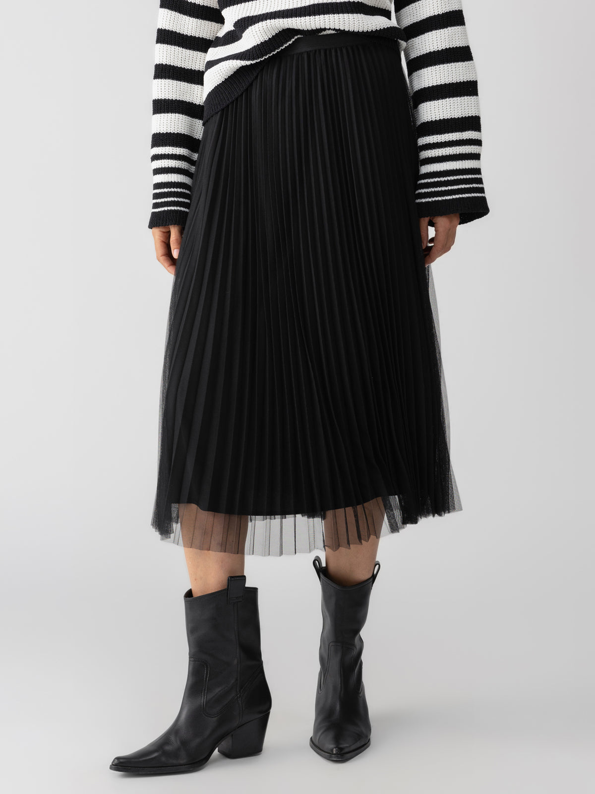 A person is wearing a Sanctuary Clothing Tulle Pleated Midi Skirt in Black, black mid-calf heeled boots, and a long-sleeved black and white striped sweater. The photo is taken from the shoulders down against a plain background.