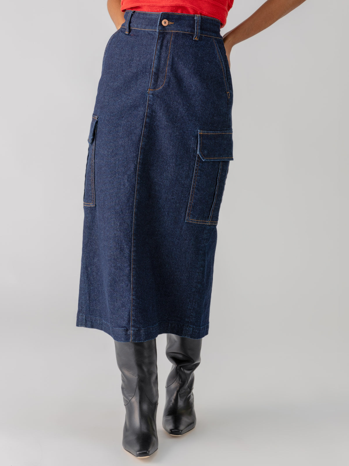 A person wearing the Sanctuary Clothing Essential Cargo Skirt Magnetic, a long dark denim skirt with large pockets on the sides. The skirt extends below the knees and is paired with a red top and tall black boots, against a plain, light-colored background.