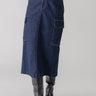 A person wearing the Sanctuary Clothing Essential Cargo Skirt Magnetic, a long dark denim skirt with large pockets on the sides. The skirt extends below the knees and is paired with a red top and tall black boots, against a plain, light-colored background.