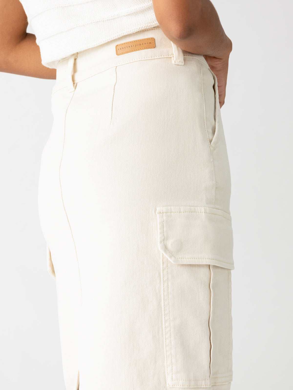 A person is wearing the Essential Cargo Skirt in French Vanilla by Sanctuary Clothing, which features a high waist and a side pocket on the right leg. The skirt appears to be made from a denim-like fabric. The person’s hand rests in their pocket, and a small leather patch with an embossed logo is visible near the waistband.