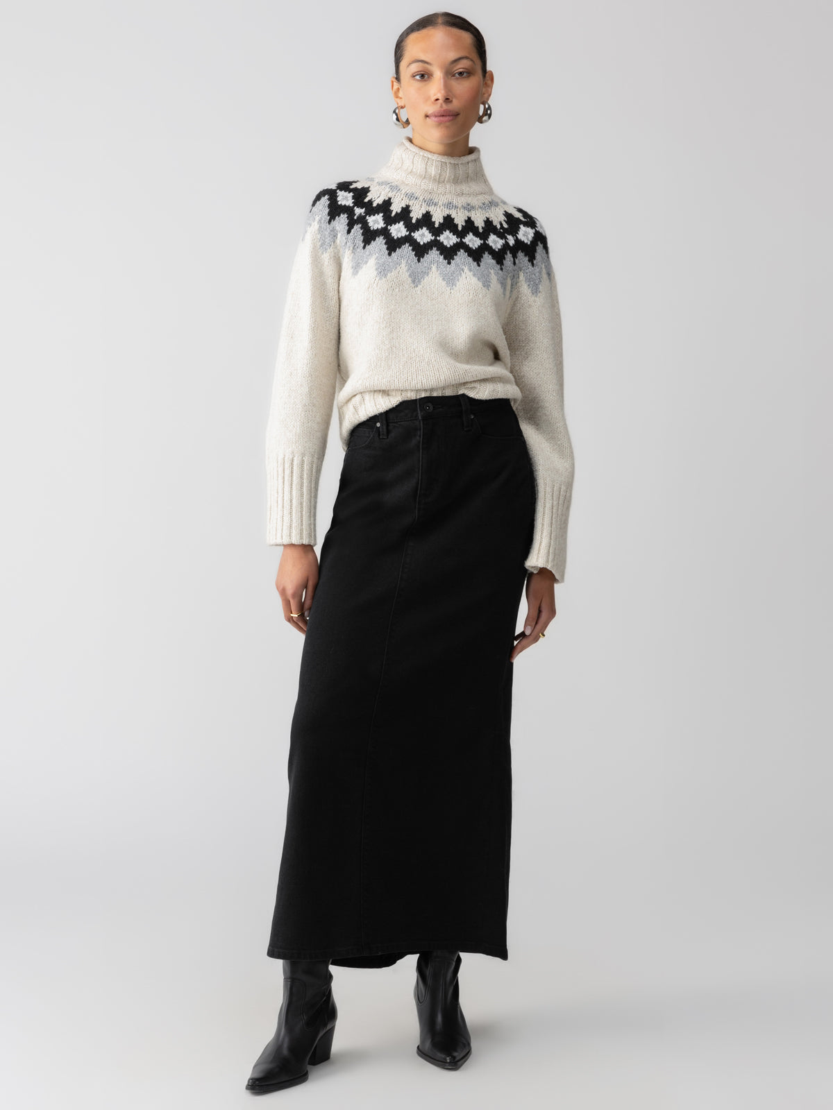 A person stands against a plain background, wearing a white turtleneck sweater with a black and gray geometric pattern, the Denim Maxi Skirt in Vintage Black by Sanctuary Clothing, black boots, and hoop earrings. The person has their hair pulled back and poses with one hand in their pocket.