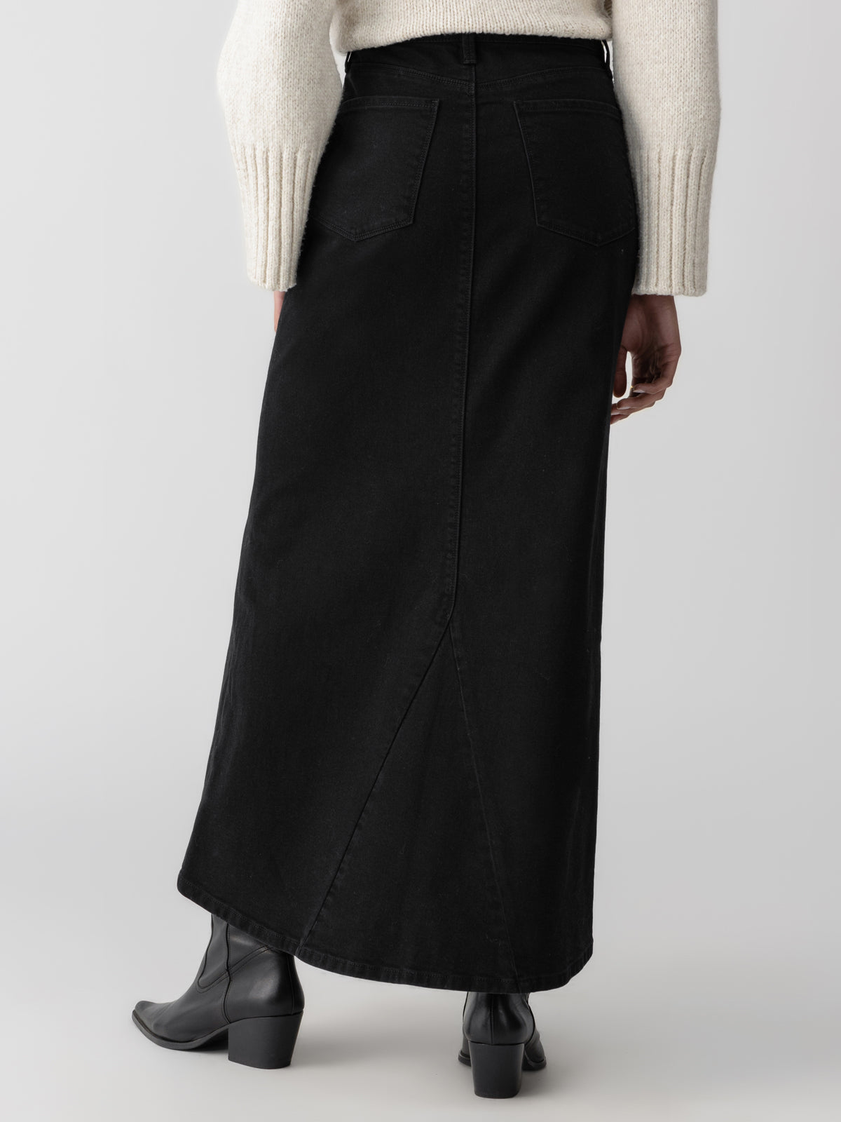 A person wearing the Sanctuary Clothing Denim Maxi Skirt in Vintage Black and a cream-colored sweater stands facing away from the camera. The skirt features back pockets and a seam down the center. The individual is also wearing black ankle boots with a low heel.