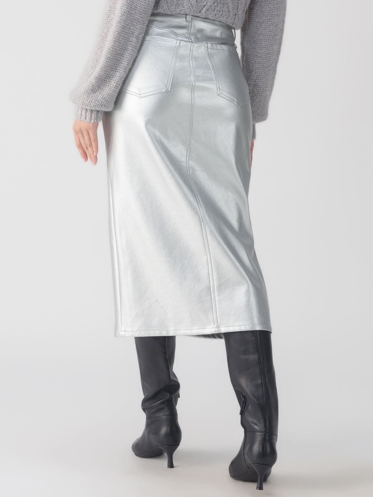 A person wearing Sanctuary Clothing's leather-like midi skirt in silver, featuring a back slit, and paired with black knee-high boots. The ensemble is completed with a gray textured knit sweater, set against a plain light gray background.