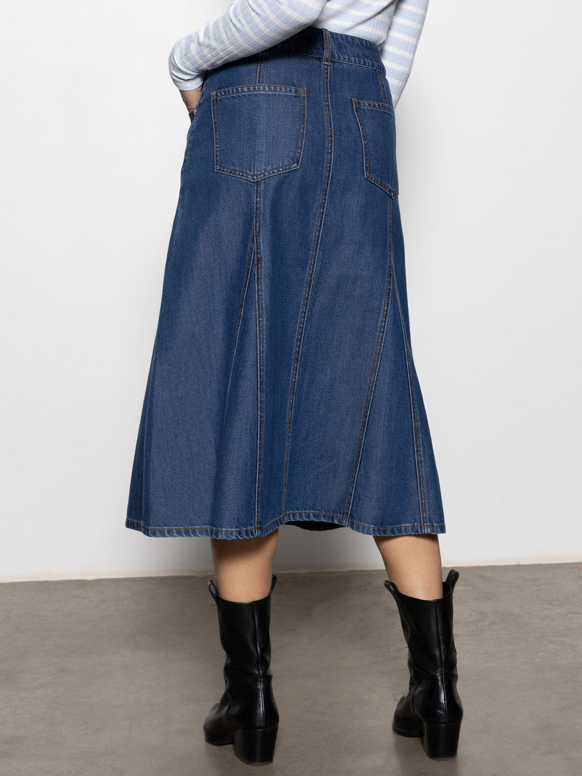 A person wearing the "Flora Denim Skirt Happy Hour" by Sanctuary Clothing, paired with black boots. Their top is light blue with white stripes, and they stand on a light gray floor against a plain white background.