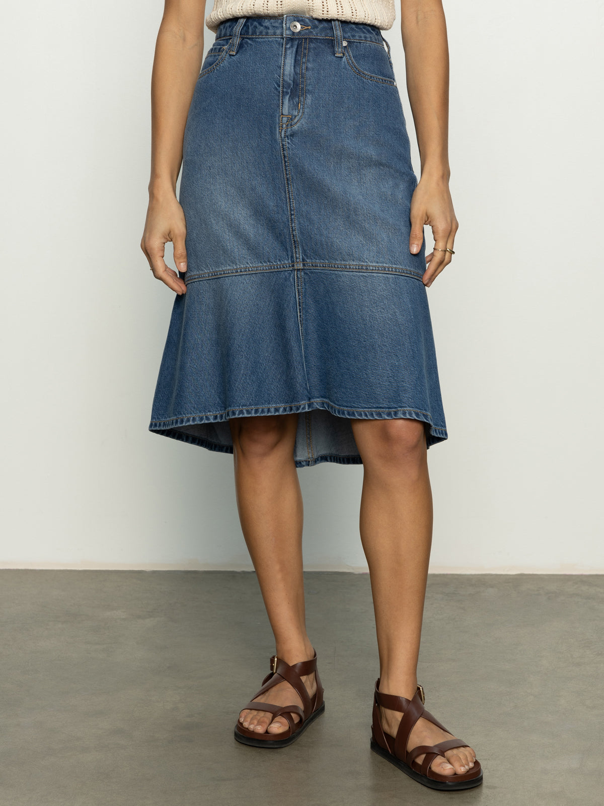 A person wearing Sanctuary Clothing's Piper Skirt Love Fool, a light top, and brown leather sandals stands against a plain background. The skirt is blue denim with a high-low hem. Their hands rest by their sides.