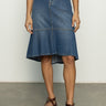 A person wearing Sanctuary Clothing's Piper Skirt Love Fool, a light top, and brown leather sandals stands against a plain background. The skirt is blue denim with a high-low hem. Their hands rest by their sides.