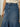 A person is wearing the Piper Skirt Love Fool by Sanctuary Clothing, featuring a blue denim design with contrast stitching, white button, flared bottom, and two front pockets. They are also donning a cream-colored sleeveless knit top against a plain background.