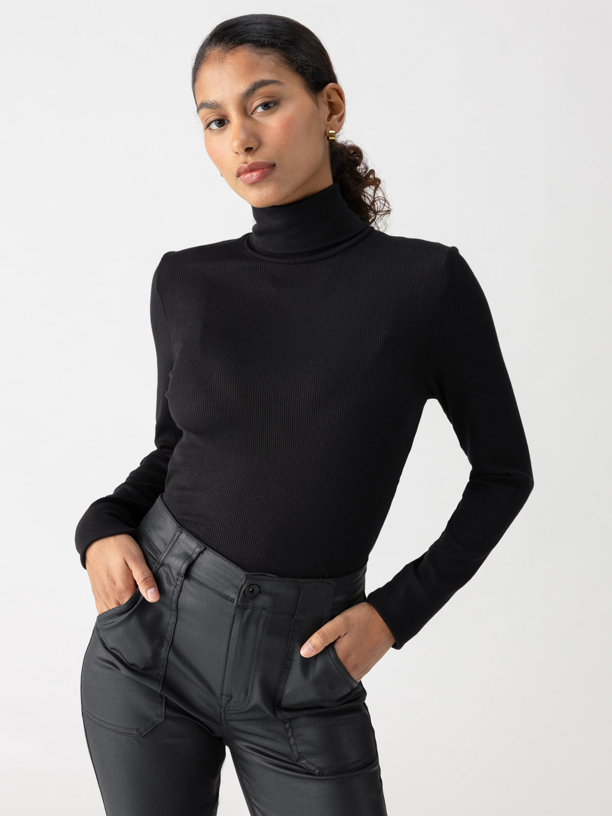 A person with dark, curly hair pulled back is wearing the Essential Turtleneck Top Black by Sanctuary Clothing and black high-waisted pants. They stand with hands in pockets, looking confidently at the camera against a plain light background.